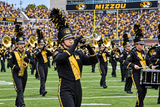A marching band performs on a football field in a stadium, with a large crowd in the background. The band members wear black and gold uniforms with black and gold plumed hats and play brass and percussion instruments.