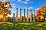 A scenic outdoor photograph of six tall stone columns on a grassy lawn, with a domed statehouse in the background and autumn trees in full color under a blue sky with sunlight filtering through the leaves.